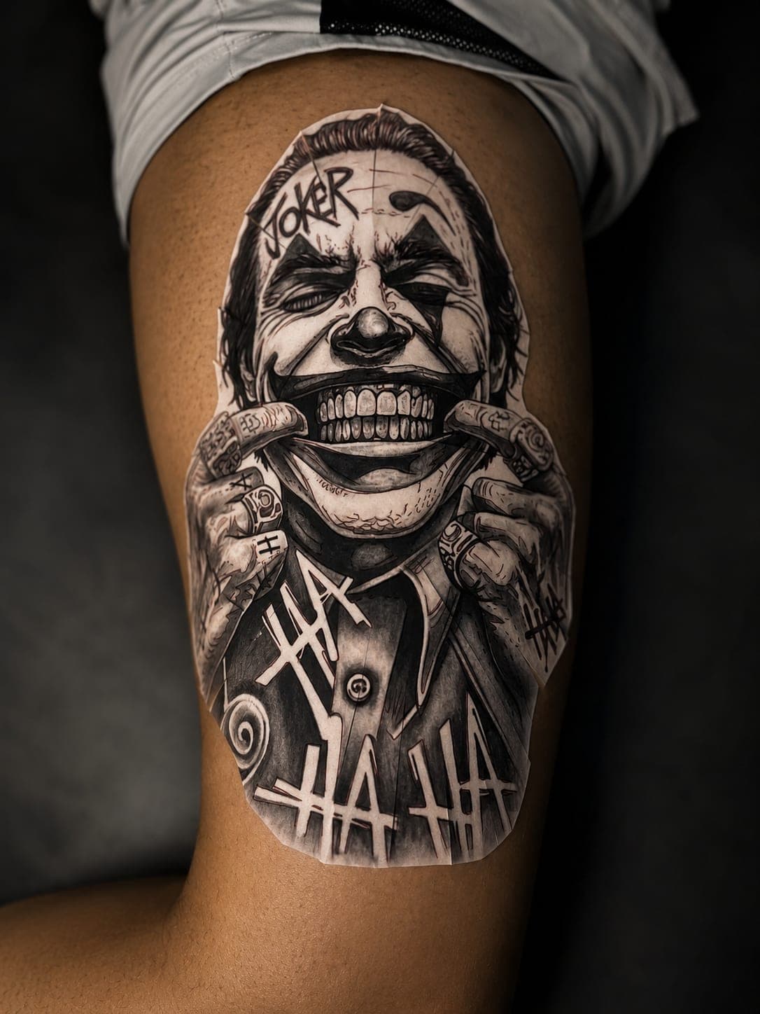 Retrato del Joker en realismo black and grey sobre el muslo, con lettering HA HA a los lados